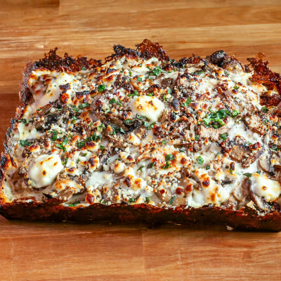 Close-up of a rectangular mushroom and cheese flatbread, garnished with fresh herbs, on a wooden surface.