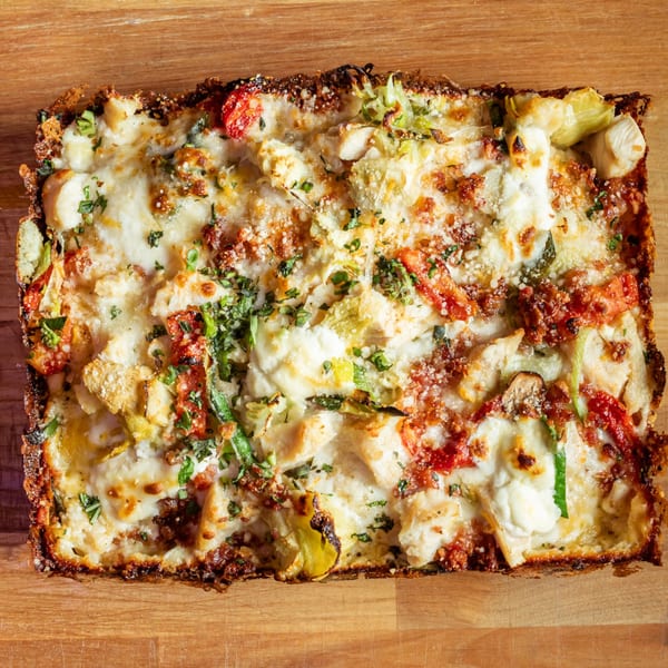 A rectangular Detroit-style pizza with melted cheese, chicken, tomatoes, and green herbs on a wooden surface.