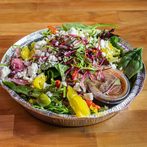 A vibrant salad with mixed greens, feta, peppers, and dressing in a foil takeout container.