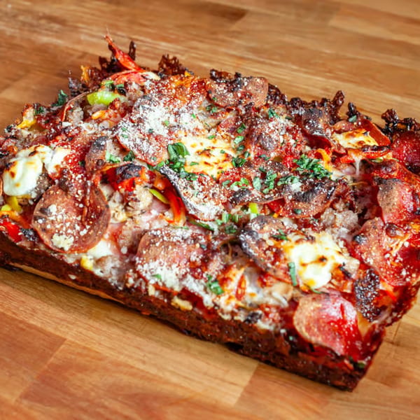 A rectangular Detroit-style pizza slice with pepperoni, sausage, peppers, and melted cheese on a wooden surface.