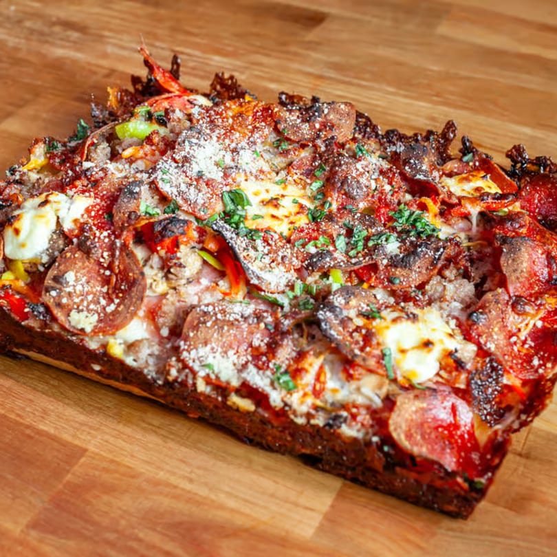 A rectangular Detroit-style pizza slice with pepperoni, sausage, peppers, and melted cheese on a wooden surface.