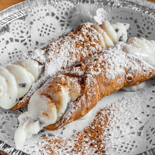 Two cannolis filled with white cream, dusted with powdered sugar and cinnamon, on a white doily.